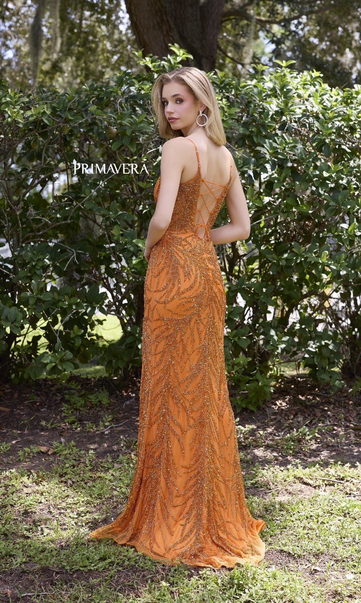 A woman poses outdoors before green bushes, wearing the Primavera 4310 Scoop-Neck Long Beaded Prom Dress featuring a corset-style bodice and lace-up back.